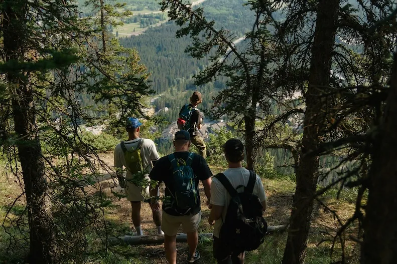A group of hikers descending a forested trail with a wide mountain valley opening below them.